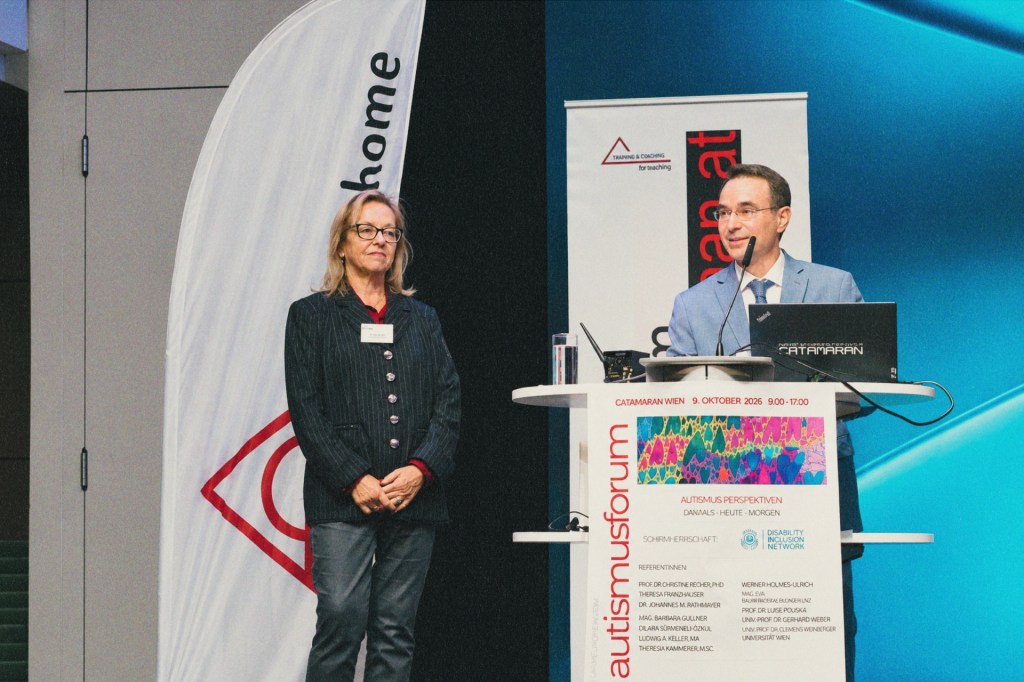 A man in a suit speaks at a lectern with the Autismusforum logo. A woman stands next to him in front of Rainman’s Home banners.