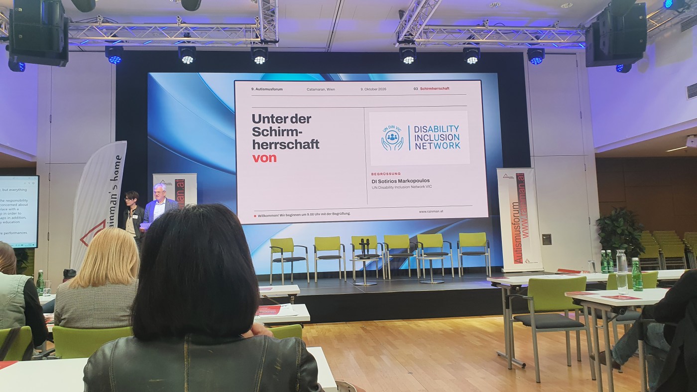Conference stage with a large screen reading “Unter der Schirmherrschaft von” next to the UN DIN VIC Disability Inclusion Network logo, with a row of green chairs on the stage.