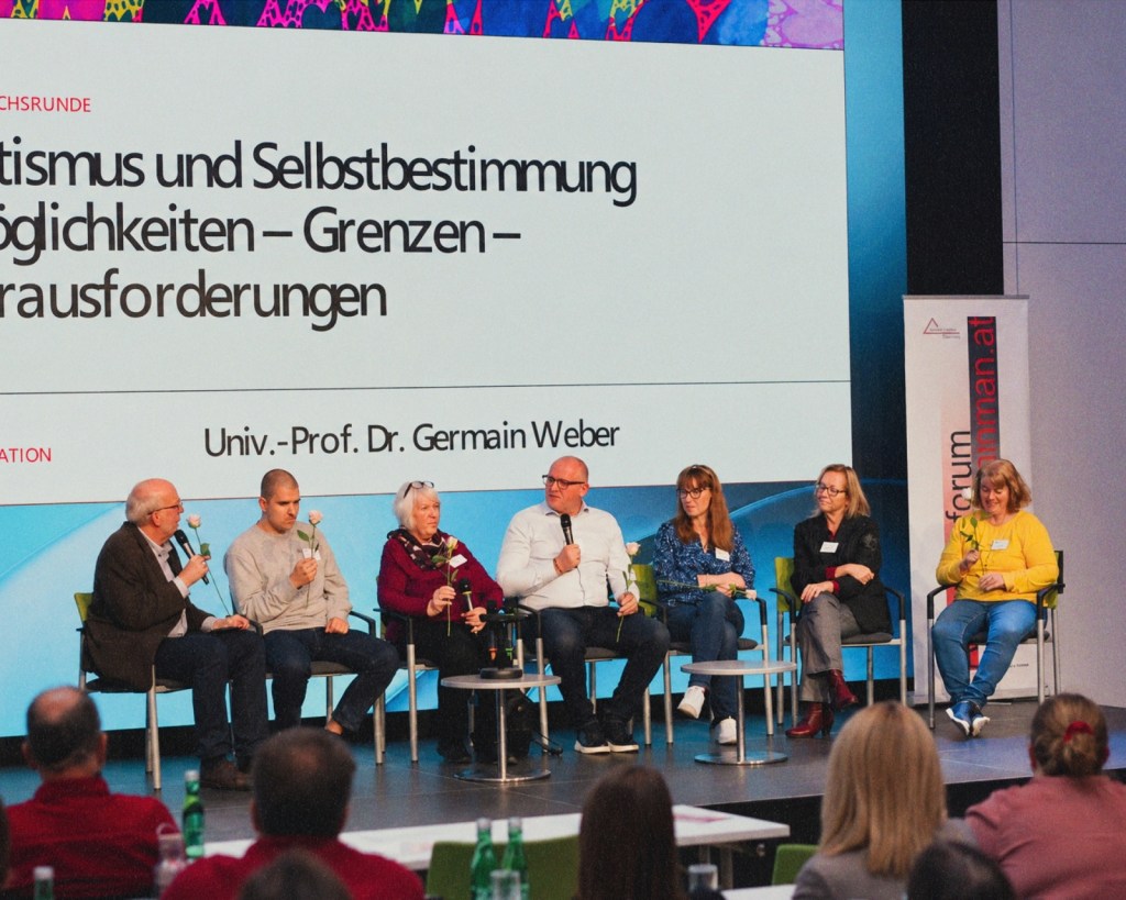 Seven people sit in a row of chairs on a stage, several holding a single white rose, two speaking into microphones. Behind them, a large screen shows the panel title on autism and self-determination and the moderator’s name, Univ.-Prof. Dr. Germain Weber. Audience members sit in the foreground.