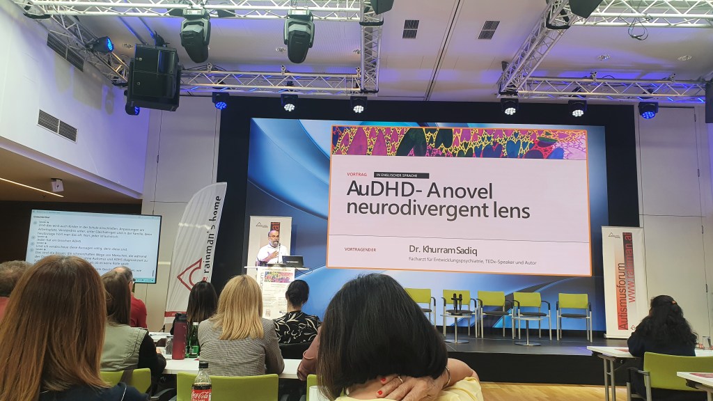 A speaker at a lectern in front of a large screen showing the title “AuDHD – A novel neurodivergent lens” and the name Dr. Khurram Sadiq, with an audience seated in the foreground and live captions on a side screen.